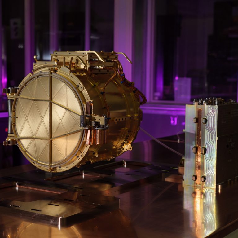 The gold colored SUDA sensor head is shown resting on a table in a clean room, seen in the center left of this image. The cover to the senor head is closed, and to the right a small, silver electronics box is visible. Combined, the pieces make up the entire SUDA instrument. The background space in the clean room is lit up purple.