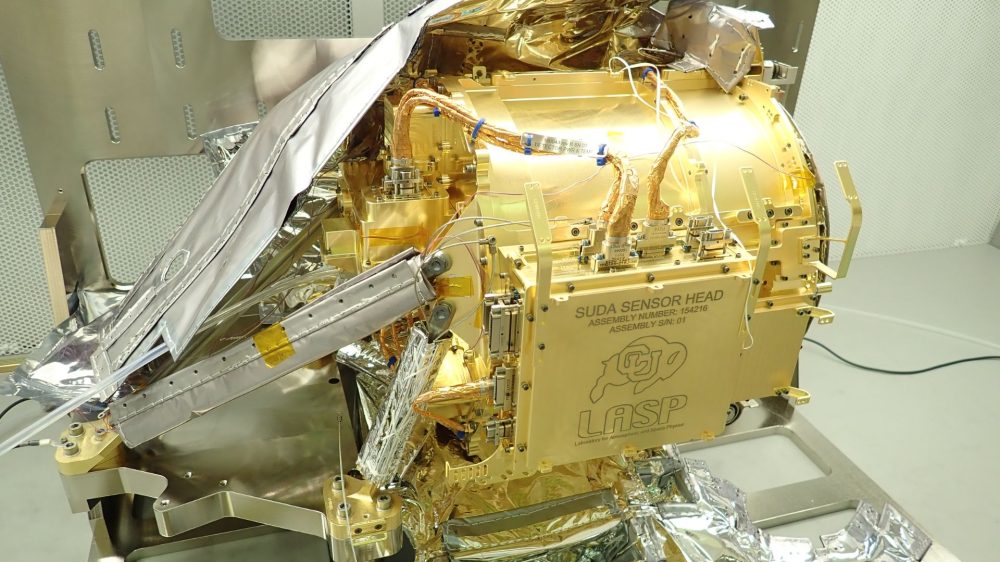 The instrument sensor head is shown in the middle of the image as a large metallic device, gold in color, with wiring attached. The sensor is resting on a table in a cleanroom. There are no people visible in the image. Multi-layer insulation is visible on the top and underneath the instrument. On the side of the instrument is a panel with information etched in, including “SUDA SENSOR HEAD”, “LASP” (which refers to the Laboratory for Atmospheric and Space Physics), and the mascot symbol for the University of Boulder, which is a buffalo.