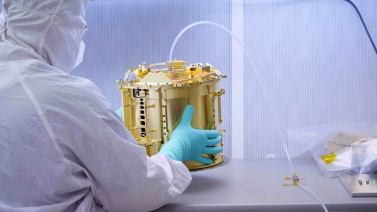 An engineer clad in a white suit holds part of the gold-plated SUrface Dust Analyzer instrument for the Europa Clipper mission