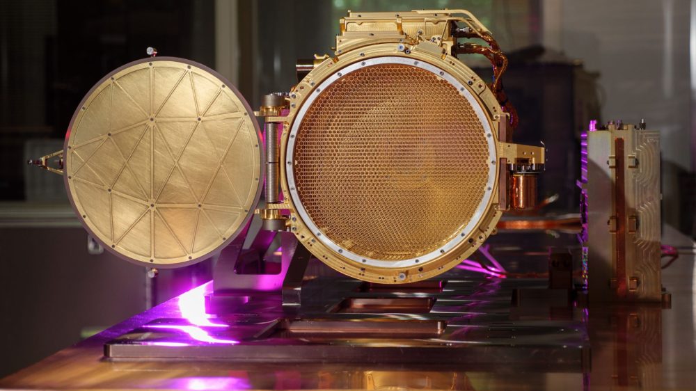The gold colored SUDA sensor head is shown resting on a table in a clean room, seen in the center of this image. The cover to the image is shown open in the image, allowing you to see a mesh screen inside the sensor head. A purple light is seen reflecting off part of the sensor head and the table it rests on.